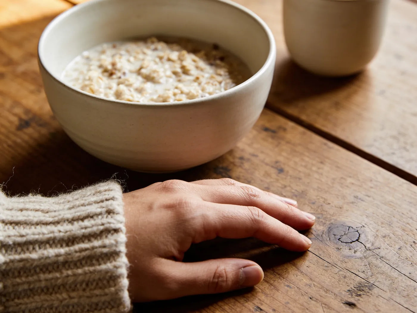 A hand resting on a worn wooden kitchen counter beside a bowl of porridge and a cup of black coffee in morning light